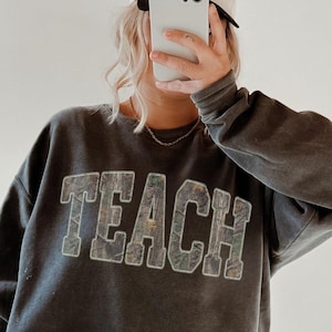 May include: A gray sweatshirt with the word "TEACH" in large, textured letters. The person is wearing black leggings, a white baseball cap, and brown clogs. The cap has text on it.