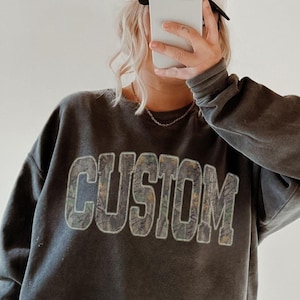 K&ouml;nnte beinhalten: Ein graues Sweatshirt mit dem Wort "CUSTOM" in einer verwaschenen, strukturierten Schriftart. Die Person tr&auml;gt schwarze Leggings, eine wei&szlig;e Baseballkappe und braune Hausschuhe. Das Bild wird vor einem Spiegel aufgenommen.