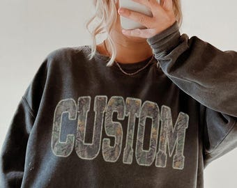 May include: A gray sweatshirt with the word "CUSTOM" in a distressed, textured font. The person is wearing black leggings, a white baseball cap, and brown slippers. The image is taken in front of a mirror.