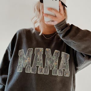 May include: Dark gray sweatshirt with the word "MAMA" in large, textured letters across the front. The letters have a floral pattern. The person is wearing black leggings and a white baseball cap.