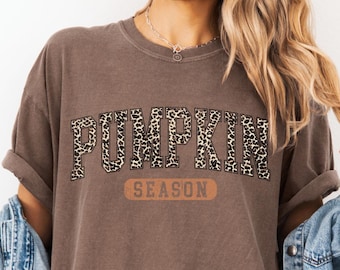 May include: A brown t-shirt with the words "PUMPKIN SEASON" printed on the front. The word "PUMPKIN" is in a leopard print pattern, and "SEASON" is in a brown font. The shirt has rolled-up sleeves.