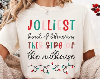 May include: Cream-colored t-shirt with the text "JOLLIEST bunch of librarians THIS SIDE of the nuthouse" in colorful letters. The shirt features a string of Christmas lights at the bottom. The person is wearing red and white plaid pajama pants and holding a Santa mug.