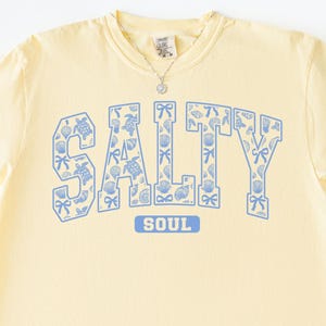 May include: A light yellow t-shirt with the word "SALTY" in blue, filled with seashell and bow graphics. The word "SOUL" is below "SALTY". A silver necklace is visible. The word "Banana" is written in cursive at the bottom right.