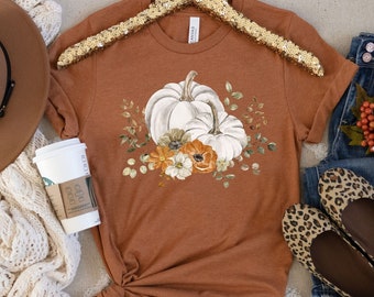 May include: A rust-colored t-shirt with a white pumpkin and floral design. The shirt is styled with a brown hat, a white coffee cup, a white crocheted scarf, blue jeans, and leopard print flats.
