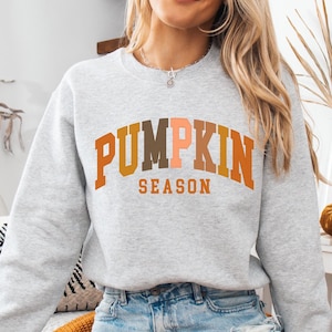May include: A light grey crew neck sweatshirt featuring the text "PUMPKIN SEASON" in orange, brown, and peach. The image has a fall theme. The sweatshirt is a casual style.