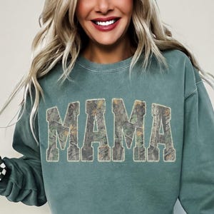 May include: A teal green sweatshirt with the word "MAMA" in large, block letters across the front. The letters have a camouflage pattern. The model is wearing light wash denim shorts and a black and white checkered scrunchie.