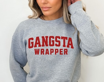 May include: A light gray sweatshirt with the words "GANSTA WRAPPER" printed in red block letters. The sweatshirt has long sleeves and a crew neck. The model is wearing denim shorts.