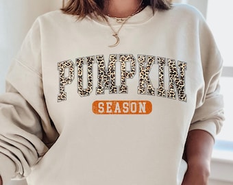 May include: Beige sweatshirt with the words "PUMPKIN SEASON" printed on the front. The word "PUMPKIN" is in a leopard print design, and "SEASON" is in an orange rectangle. The sweatshirt has long sleeves and a crew neck.