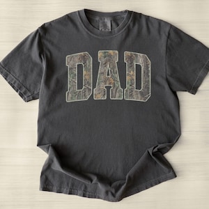 May include: Dark grey t-shirt featuring the word "DAD" in large, block letters. The letters are filled with a camouflage pattern in green, brown, and tan. The shirt has short sleeves and a crew neck.