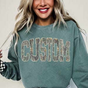 K&ouml;nnte beinhalten: Ein teal-farbenes Sweatshirt mit dem Wort "CUSTOM" in gro&szlig;en Blockbuchstaben. Die Buchstaben sind mit einem Camouflage-Muster gef&uuml;llt. Das Sweatshirt hat lange &Auml;rmel und einen Rundhalsausschnitt. Das Model tr&auml;gt Jeansshorts.