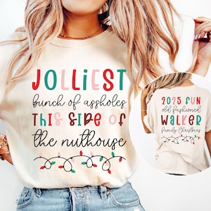 Może przedstawiać: Kremowy t-shirt z napisem "JOLLIEST bunch of assholes THIS SIDE of the nuthouse" w kolorze czerwonym, zielonym i różowym. Koszulka posiada również łańcuch lampek choinkowych. Z tyłu koszulki napis "2025 FUN old fashioned Walker Family Christmas".