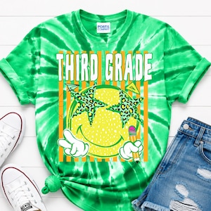 May include: A green tie-dye t-shirt featuring the text THIRD GRADE in white letters. The graphic includes a yellow smiley face with leopard print star-shaped glasses, a hand making a peace sign, and a yellow pencil. White sneakers and denim shorts.