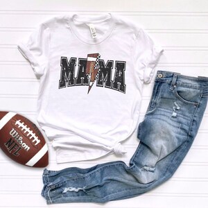 May include: White t-shirt with a black and white graphic of a football and the word "MAMA" in a distressed font. The shirt is paired with blue jeans.
