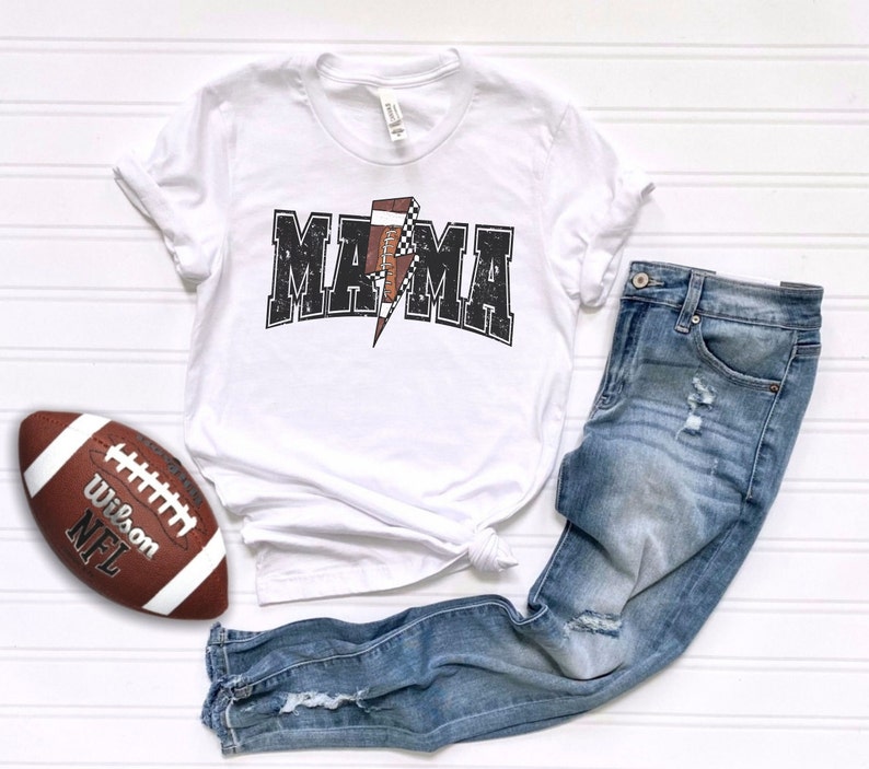 May include: White t-shirt with a black and white graphic of a football and the word "MAMA" in a distressed font. The shirt is paired with blue jeans.