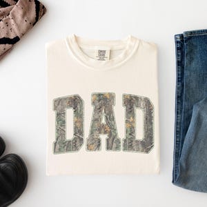 May include: A cream-colored short-sleeve t-shirt featuring the word DAD printed in large, block-style letters with a green, brown, and tan camouflage pattern. The shirt has a crew neckline and a Comfort Colors brand label at the inner collar.