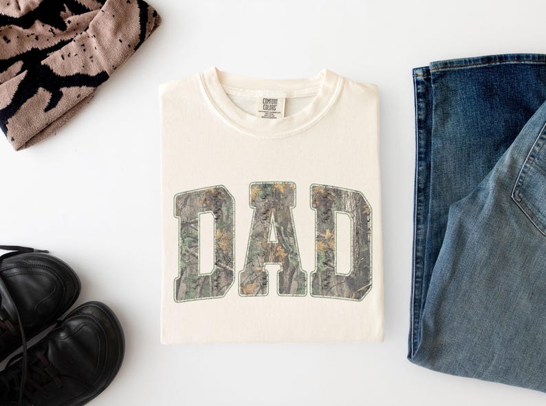 May include: A cream-colored short-sleeve t-shirt featuring the word DAD printed in large, block-style letters with a green, brown, and tan camouflage pattern. The shirt has a crew neckline and a Comfort Colors brand label at the inner collar.
