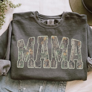 May include: Dark gray sweatshirt with the word "MAMA" in large, block letters. The letters are filled with a camouflage pattern and outlined in white. The sweatshirt has a crew neck and long sleeves.