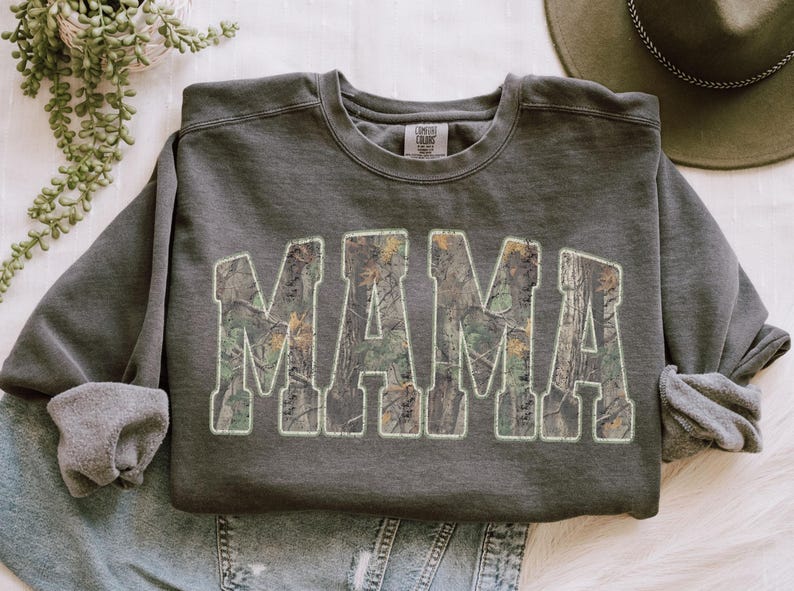 May include: Dark gray sweatshirt with the word "MAMA" in large, block letters. The letters are filled with a camouflage pattern and outlined in white. The sweatshirt has a crew neck and long sleeves.