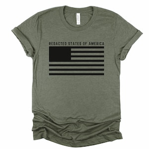 K&ouml;nnte beinhalten: Olivgr&uuml;nes T-Shirt mit schwarzer, zensierter amerikanischer Flagge und dem Text "REDACTED STATES OF AMERICA". Das Design ist auf der Vorderseite aufgedruckt. Das T-Shirt hat kurze &Auml;rmel und einen Rundhalsausschnitt.