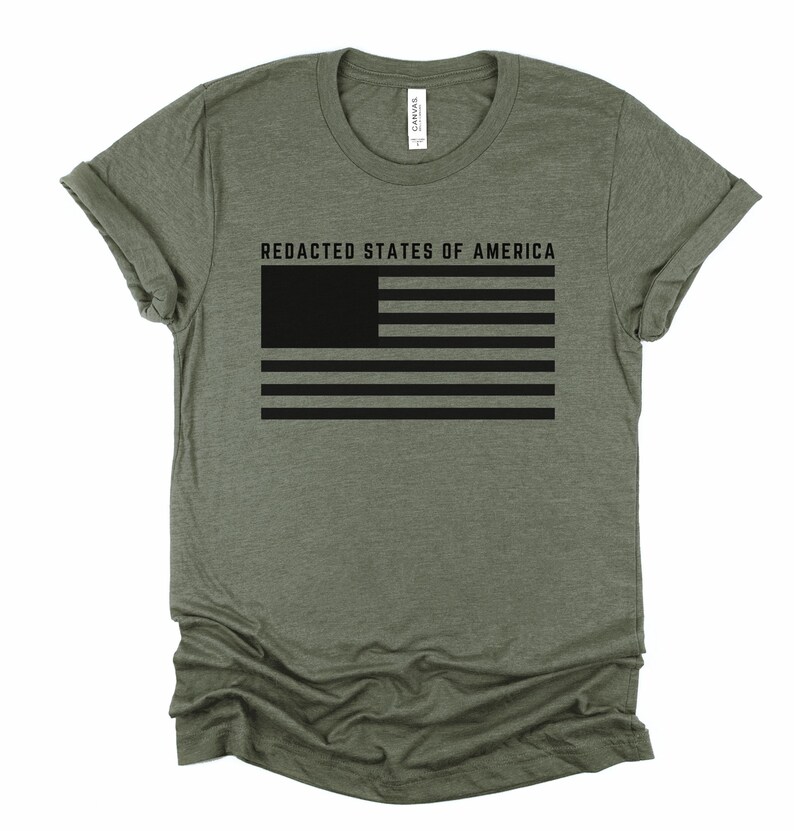 K&ouml;nnte beinhalten: Olivgr&uuml;nes T-Shirt mit schwarzer, zensierter amerikanischer Flagge und dem Text "REDACTED STATES OF AMERICA". Das Design ist auf der Vorderseite aufgedruckt. Das T-Shirt hat kurze &Auml;rmel und einen Rundhalsausschnitt.
