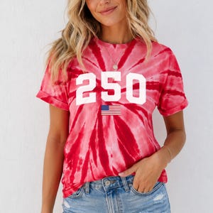 May include: Red tie-dye t-shirt featuring the number "250" in white and a small American flag graphic. The shirt is worn with denim shorts. The style is casual and summery.