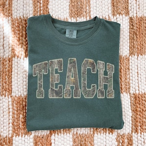 May include: A teal Comfort Colors t-shirt with the word "TEACH" in large, block letters across the front. The letters have a camouflage pattern fill and a white outline. The shirt is folded on a textured, neutral-toned surface.