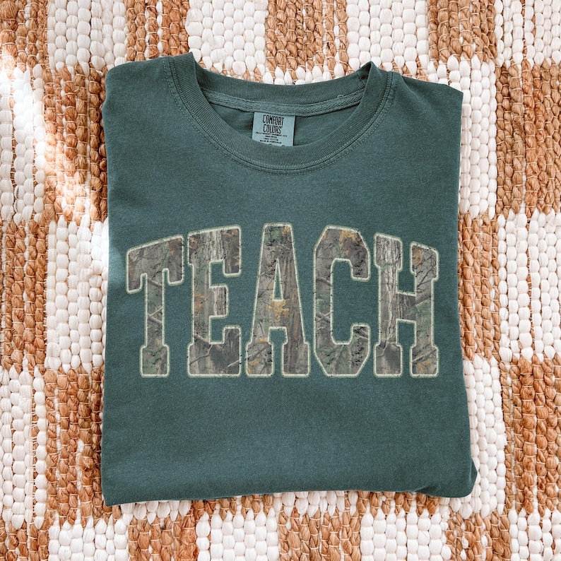 May include: A teal Comfort Colors t-shirt with the word "TEACH" in large, block letters across the front. The letters have a camouflage pattern fill and a white outline. The shirt is folded on a textured, neutral-toned surface.