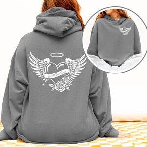 May include: A gray hoodie with a white graphic on the back featuring a heart with wings, a halo, and roses. The text "Loved One Here" is printed on a banner across the heart. A smaller version of the graphic is on the front.