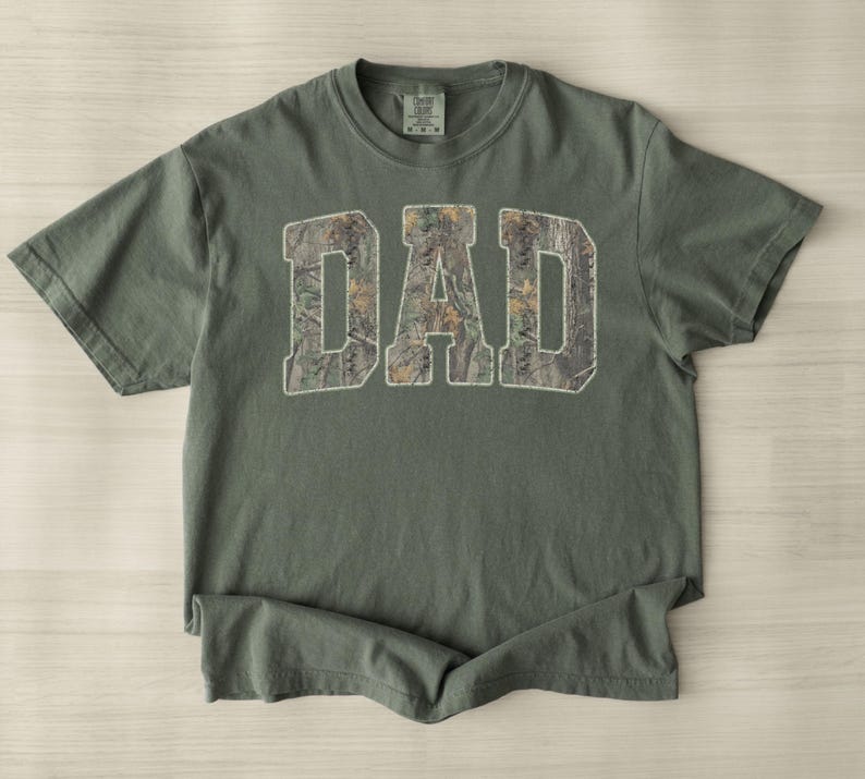 May include: A sage green short-sleeve t-shirt featuring the word DAD printed in large block letters across the chest. The letters are filled with a brown and green camouflage pattern. The shirt has a crew neckline and a standard fit.