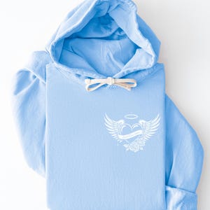 May include: A light blue hoodie with a white graphic on the front. The graphic features a heart with wings, a halo, and roses, with the text "Good Kind Soul". The hoodie has a drawstring and a folded cuff.