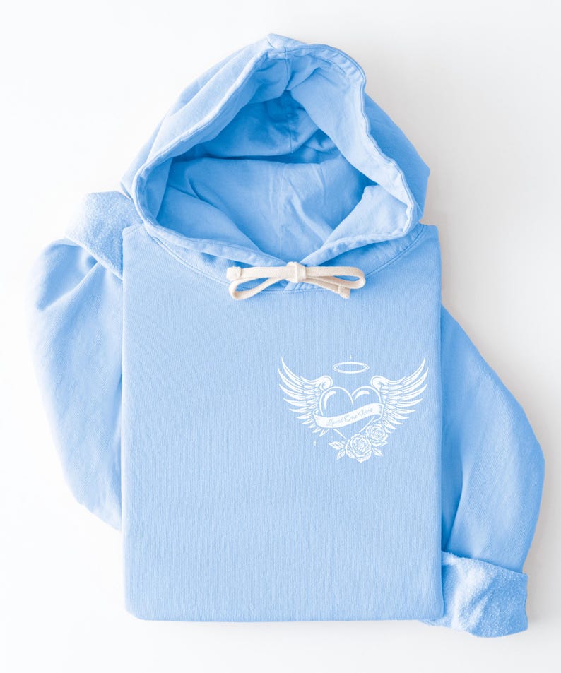 May include: A light blue hoodie with a white graphic on the front. The graphic features a heart with wings, a halo, and roses, with the text "Good Kind Soul". The hoodie has a drawstring and a folded cuff.