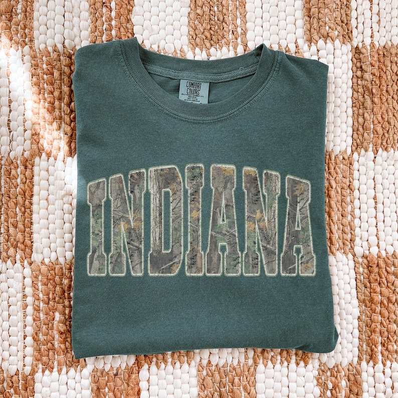 K&ouml;nnte beinhalten: Ein dunkel petrolfarbenes T-Shirt mit dem Wort "INDIANA" in einem Camouflage-Muster auf der Vorderseite. Das Shirt ist ordentlich auf einer strukturierten, neutral get&ouml;nten Oberfl&auml;che gefaltet. Es hat einen Rundhalsausschnitt und kurze &Auml;rmel.