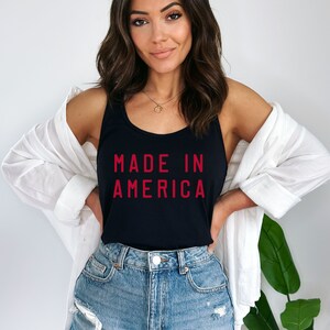 K&ouml;nnte beinhalten: Ein schwarzes Tanktop mit dem Text "MADE IN AMERICA" in roten Buchstaben. Das Model tr&auml;gt das Tanktop mit Jeans-Shorts und einem wei&szlig;en Leinenhemd.