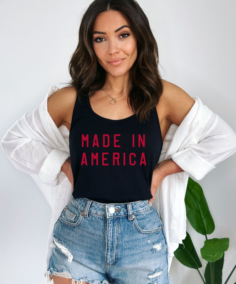 K&ouml;nnte beinhalten: Ein schwarzes Tanktop mit dem Text "MADE IN AMERICA" in roten Buchstaben. Das Model tr&auml;gt das Tanktop mit Jeans-Shorts und einem wei&szlig;en Leinenhemd.