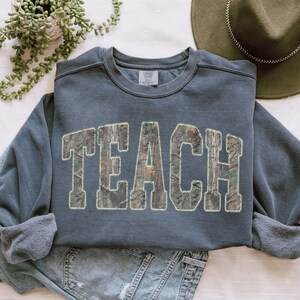 May include: A slate blue sweatshirt with the word "TEACH" in large, block letters. The letters are filled with a camouflage pattern. The sweatshirt has a crew neck and long sleeves with ribbed cuffs. A green hat and a plant are in the background.