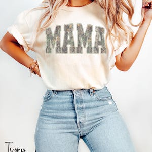 May include: Ivory-colored t-shirt with the word "MAMA" in large, camouflage-patterned letters across the chest. The shirt is paired with light blue jeans. The word "Ivory" is written in script at the bottom left.