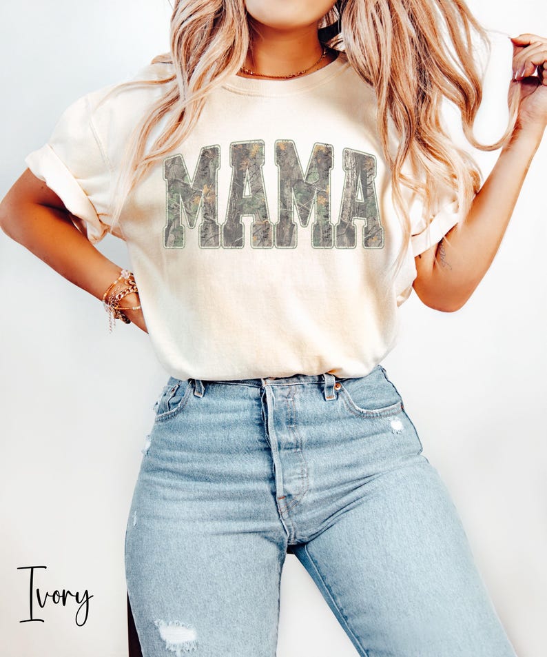 May include: Ivory-colored t-shirt with the word "MAMA" in large, camouflage-patterned letters across the chest. The shirt is paired with light blue jeans. The word "Ivory" is written in script at the bottom left.