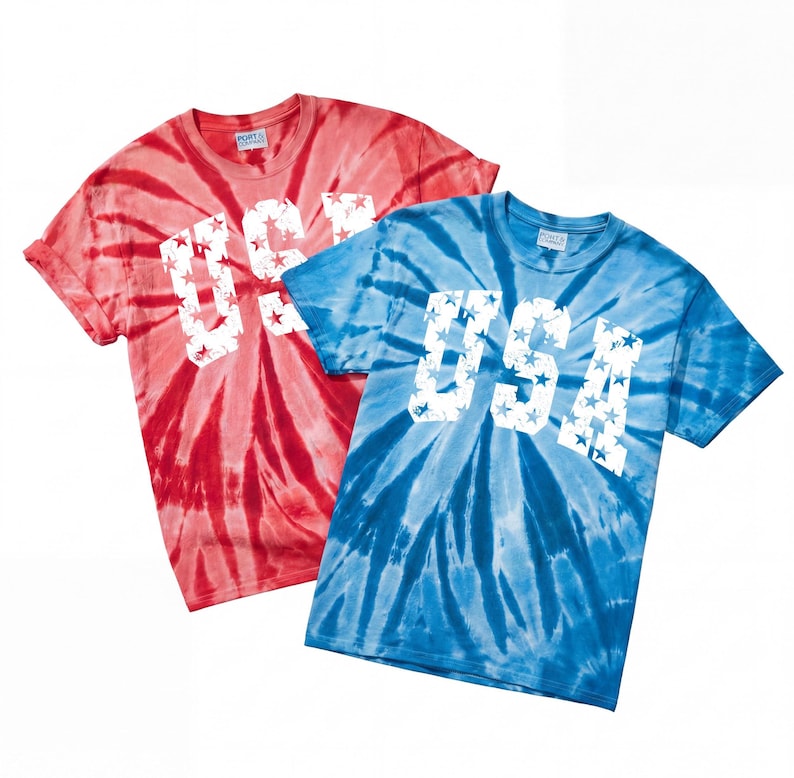May include: Two tie-dye t-shirts, one red and one blue, with the word "USA" in white block letters filled with stars. The shirts have a classic crew neck and short sleeves.