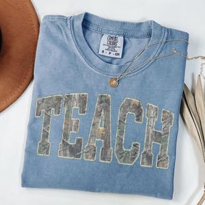 May include: A blue Comfort Colors t-shirt with the word "TEACH" in a camouflage design. A gold necklace with a pendant rests on the shirt. The shirt is folded and placed on a white surface.