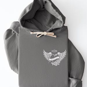 May include: A gray hoodie with a drawstring and a white graphic on the front. The graphic features a heart with wings, a halo, roses, and the text "Loved One Here."