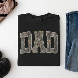 May include: A black crewneck t-shirt featuring the word DAD printed in a large, block-style font. The letters are filled with a realistic forest camouflage pattern in shades of green, brown, and tan, creating a distinct contrast against the black fabric.