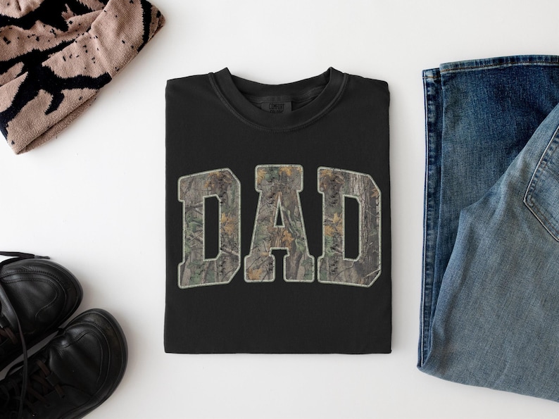 May include: A black crewneck t-shirt featuring the word DAD printed in a large, block-style font. The letters are filled with a realistic forest camouflage pattern in shades of green, brown, and tan, creating a distinct contrast against the black fabric.