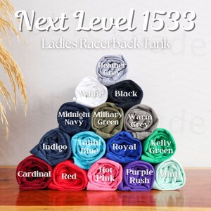 K&ouml;nnte beinhalten: Eine Auswahl an zusammengerollten T-Shirts in verschiedenen Farben, darunter Wei&szlig;, Schwarz, Marineblau, Gr&uuml;n, Grau, Blau, Rot, Rosa, Lila und Mint. Die T-Shirts sind in Pyramidenform angeordnet. Der Text "Next Level 1533 Ladies Racerback Tank" befindet sich oben im Bild.