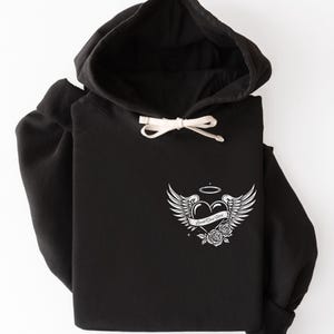 May include: Black hoodie with a drawstring hood and a white graphic on the front. The graphic features a heart with angel wings, a halo, and the words "Loved One Here" on a banner, along with roses.