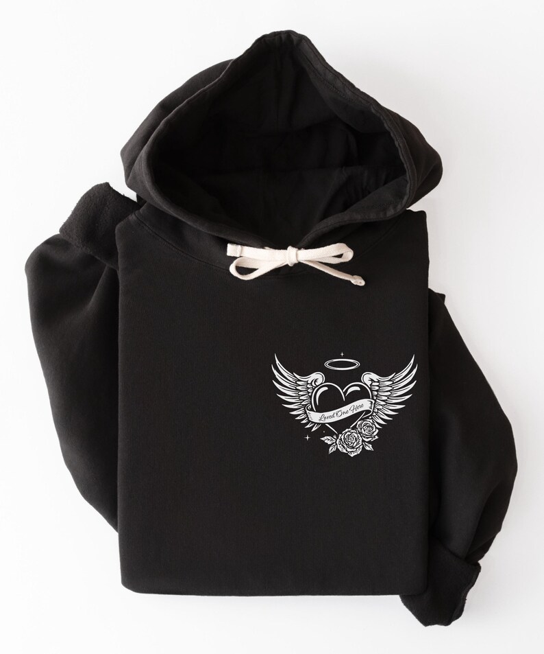 May include: Black hoodie with a drawstring hood and a white graphic on the front. The graphic features a heart with angel wings, a halo, and the words "Loved One Here" on a banner, along with roses.