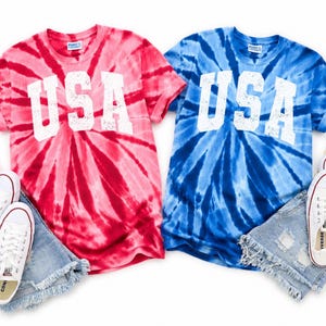 May include: Two tie-dye t-shirts, one red and one blue, each with "USA" in white block letters. The shirts are paired with white sneakers, denim shorts, and small American flags, creating a patriotic theme.