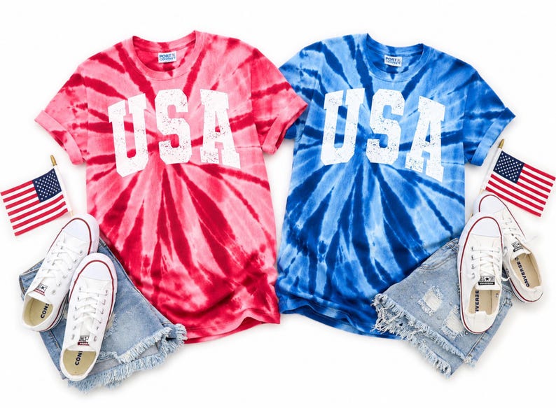 May include: Two tie-dye t-shirts, one red and one blue, each with "USA" in white block letters. The shirts are paired with white sneakers, denim shorts, and small American flags, creating a patriotic theme.