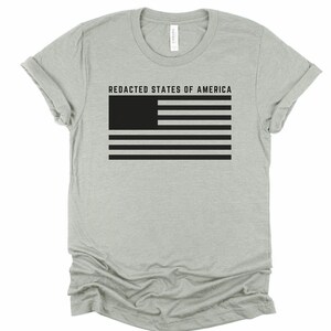K&ouml;nnte beinhalten: Hellgraues T-Shirt mit dem Text "REDACTED STATES OF AMERICA" &uuml;ber einem schwarz-wei&szlig;en Flaggen-Design. Die Flagge hat ein schwarzes Rechteck in der oberen linken Ecke und schwarze Streifen.