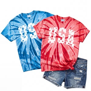 May include: Two tie-dye t-shirts, one blue and one red, with the word "USA" in white, star-filled letters. The shirts are paired with denim shorts. The shirts have a spiral pattern.