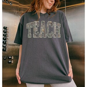 May include: Dark gray t-shirt with the word "TEACH" in large, textured letters across the front. The letters have a camouflage-like pattern. The shirt has short sleeves and a relaxed fit.